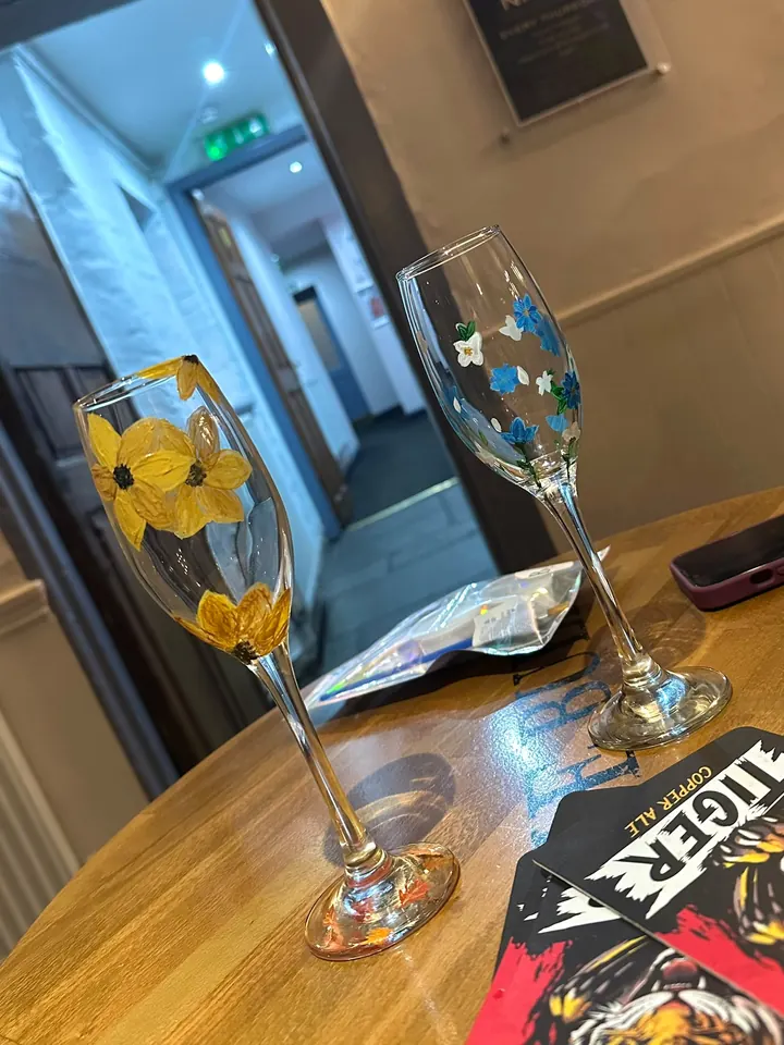Two painted champagne flutes on a pub table, one with sunflowers and one with blue and white flowers